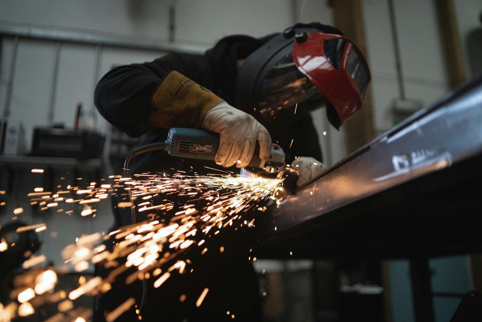 A worker in a welding helmet grinding the edge of a long steel beam, sparks trailing along it