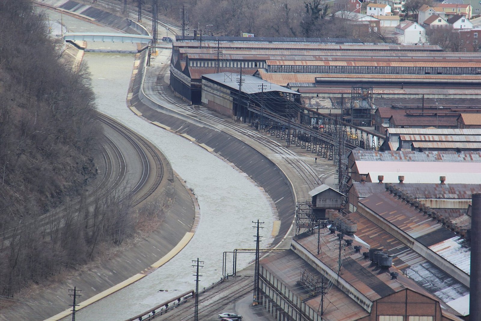 Long mill buildings with rust-coloured roofs beside a curving river channel and rail lines in Johnstown, seen from the hillside