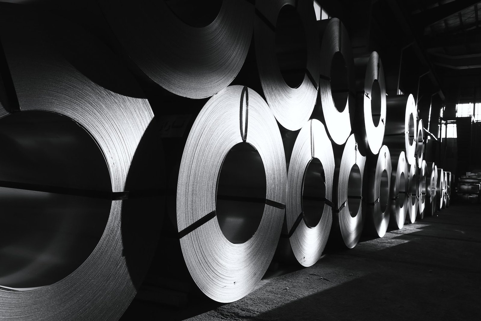 A long row of large sheet steel coils standing on edge in a dim warehouse