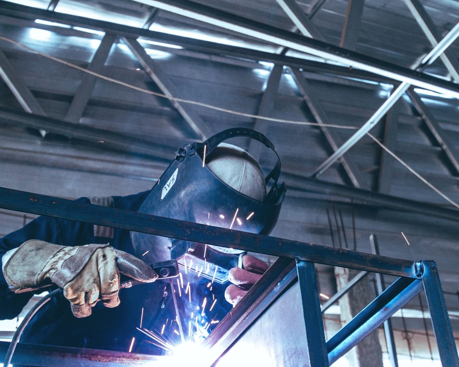 A welder in a helmet and leather gloves welding the corner of a blue steel tube frame