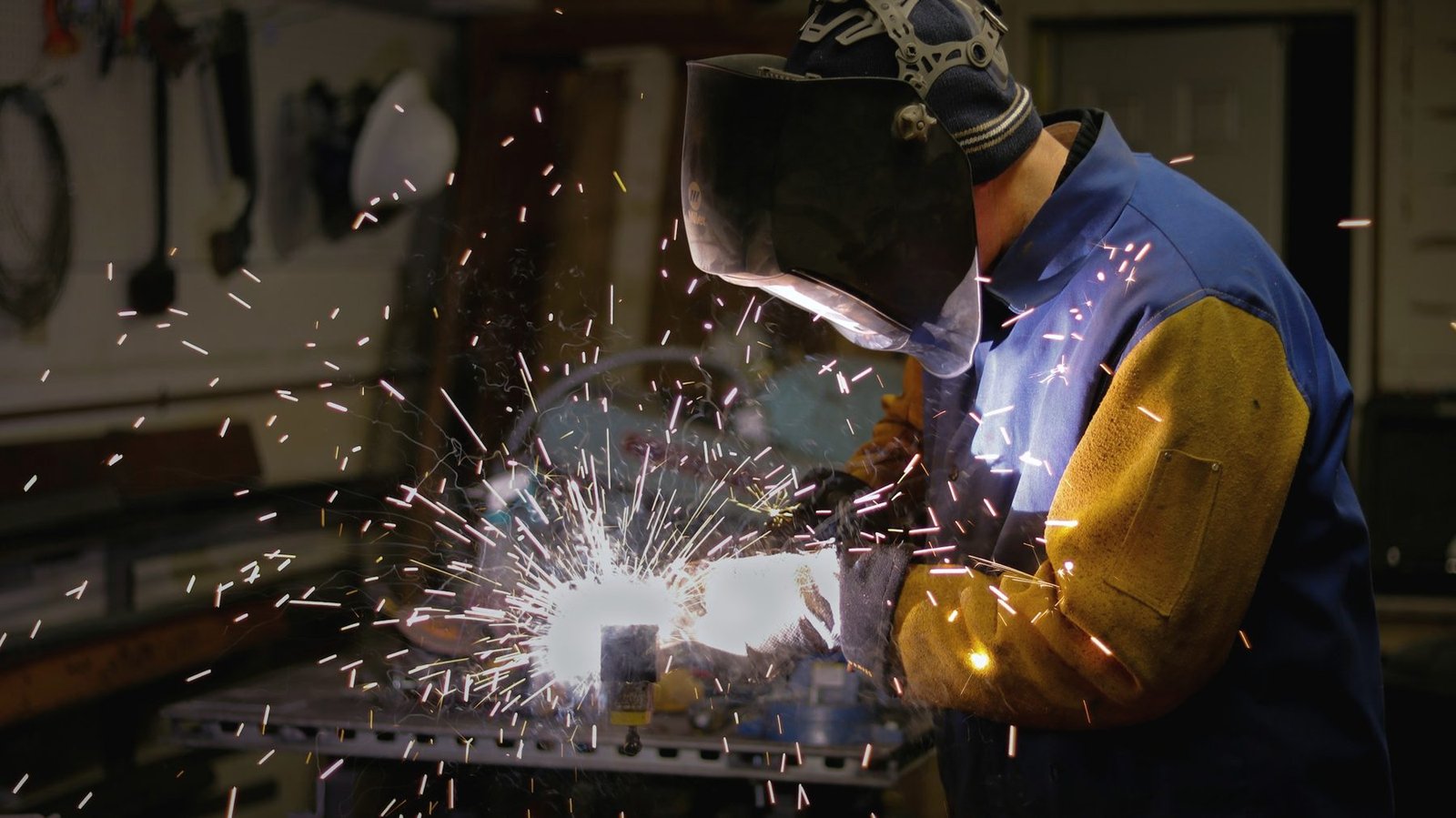 A welder in a yellow and blue jacket working at a bench in a dark workshop, sparks spreading from the weld